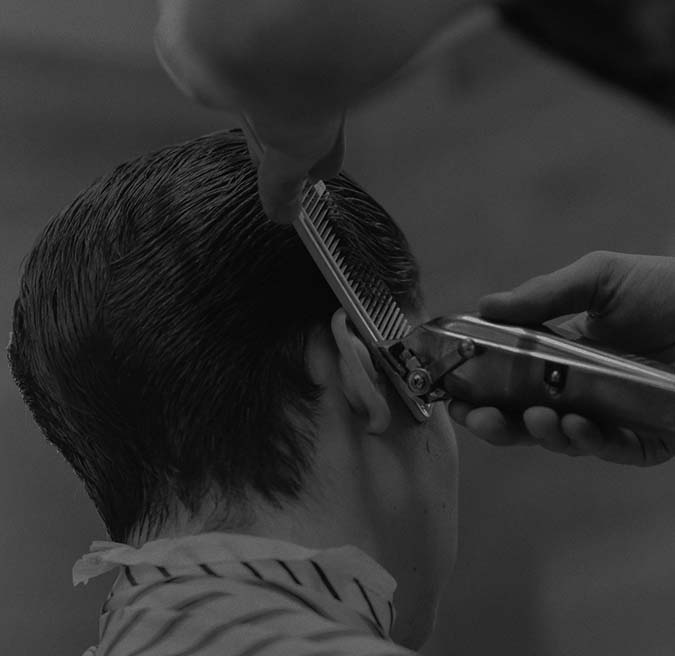 Black and white photo of a person getting a haircut with electric clippers and a comb on the back of their head.