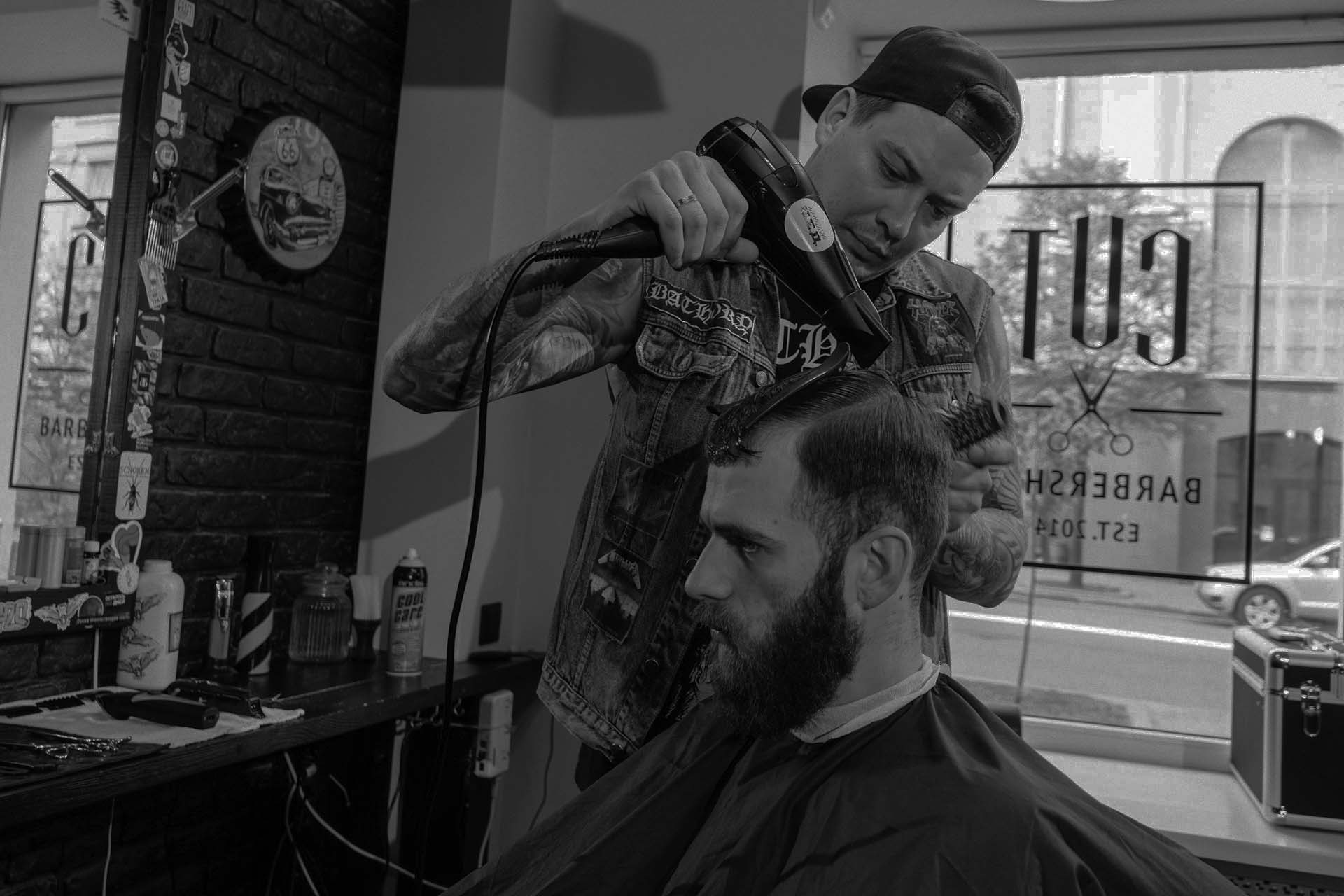 Barber using a blow dryer and comb to style a client's hair inside a vintage-style barbershop with large front window.