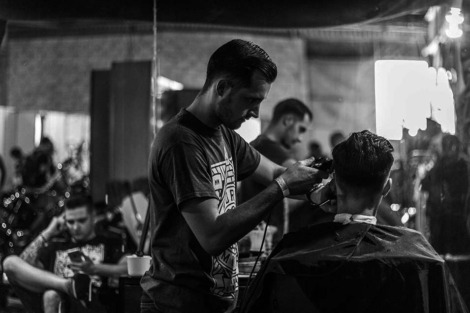 Black and white photo of a barber cutting a man's hair in a busy barbershop with mirrors and patrons in the background.